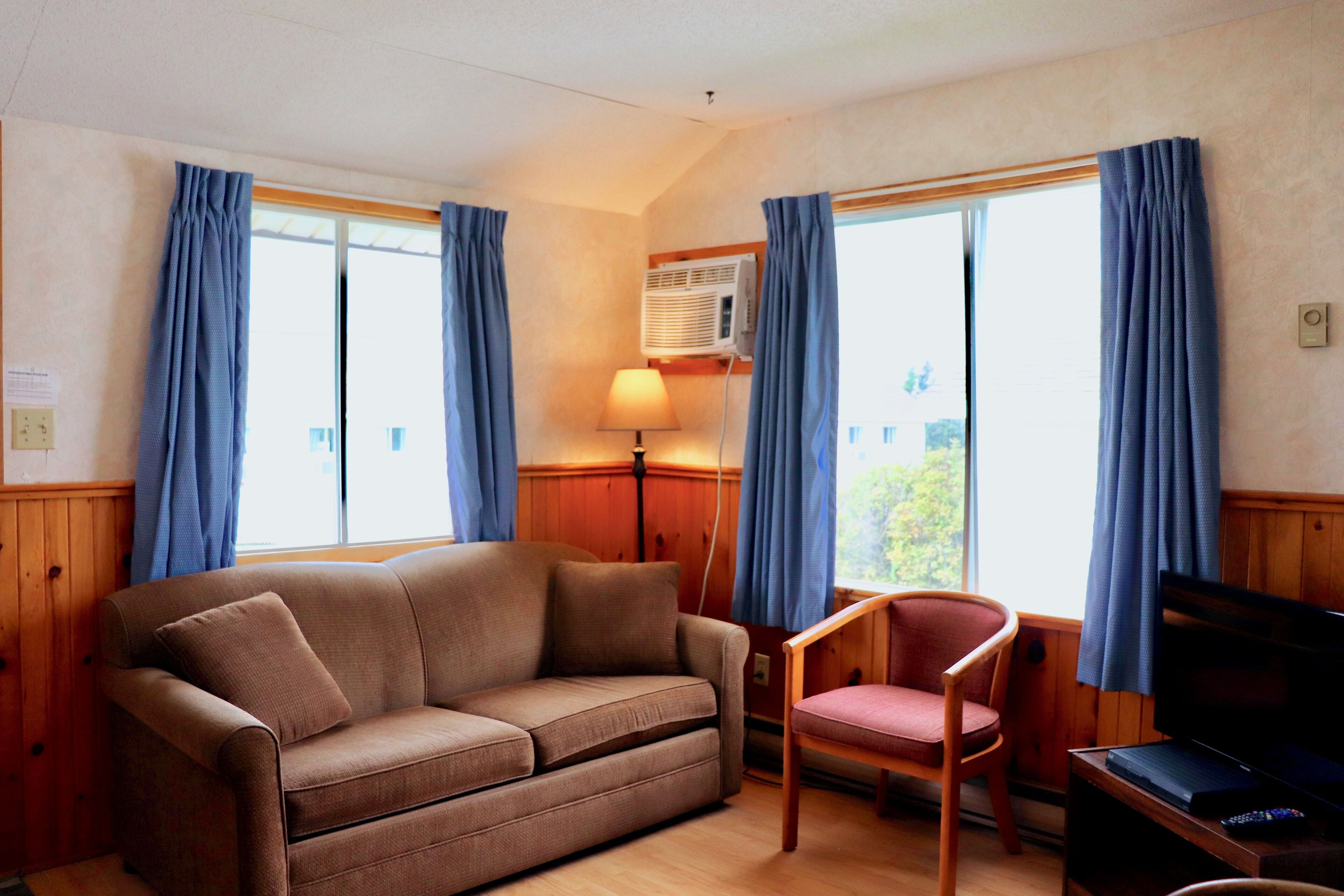 A room with a brown sofa, a wooden chair, a television, and a window with blue curtains.