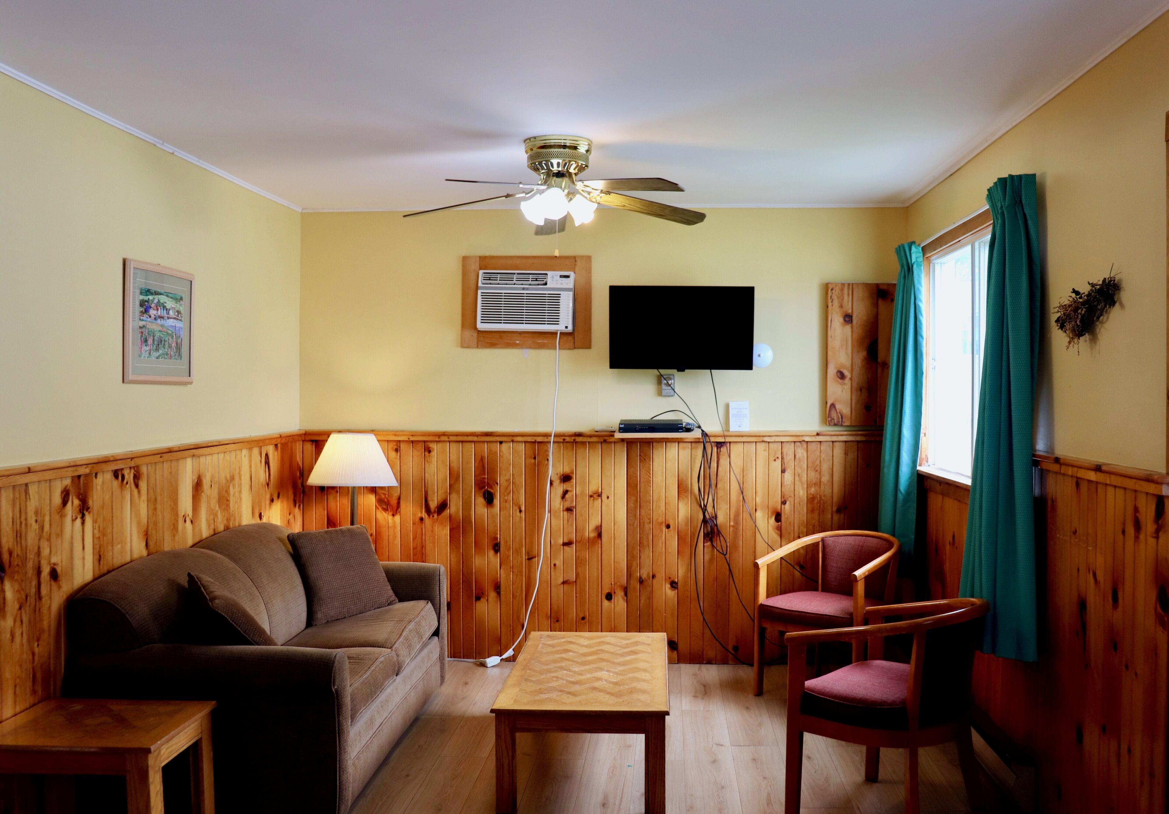 A room with a wooden paneled wall, a ceiling fan, a flat-screen TV, a wooden table, a sofa, and a window with curtains.