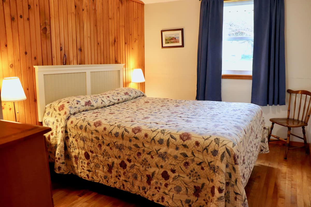 Un dormitorio con un panel de madera, una cama con ropa de cama floral, una silla y una ventana con cortinas.