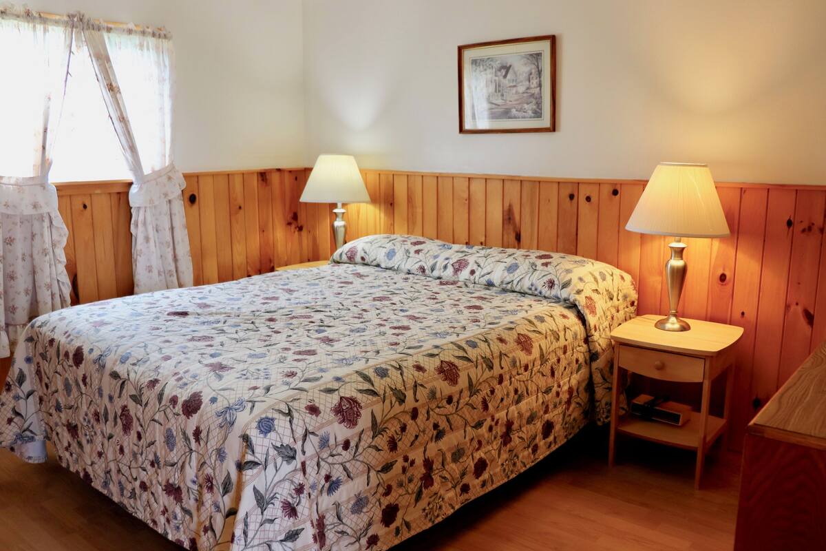 Un dormitorio con edredón estampado con motivos florales, dos lámparas de noche, un cabecero de madera, un cuadro enmarcado en la pared y una ventana con cortinas.