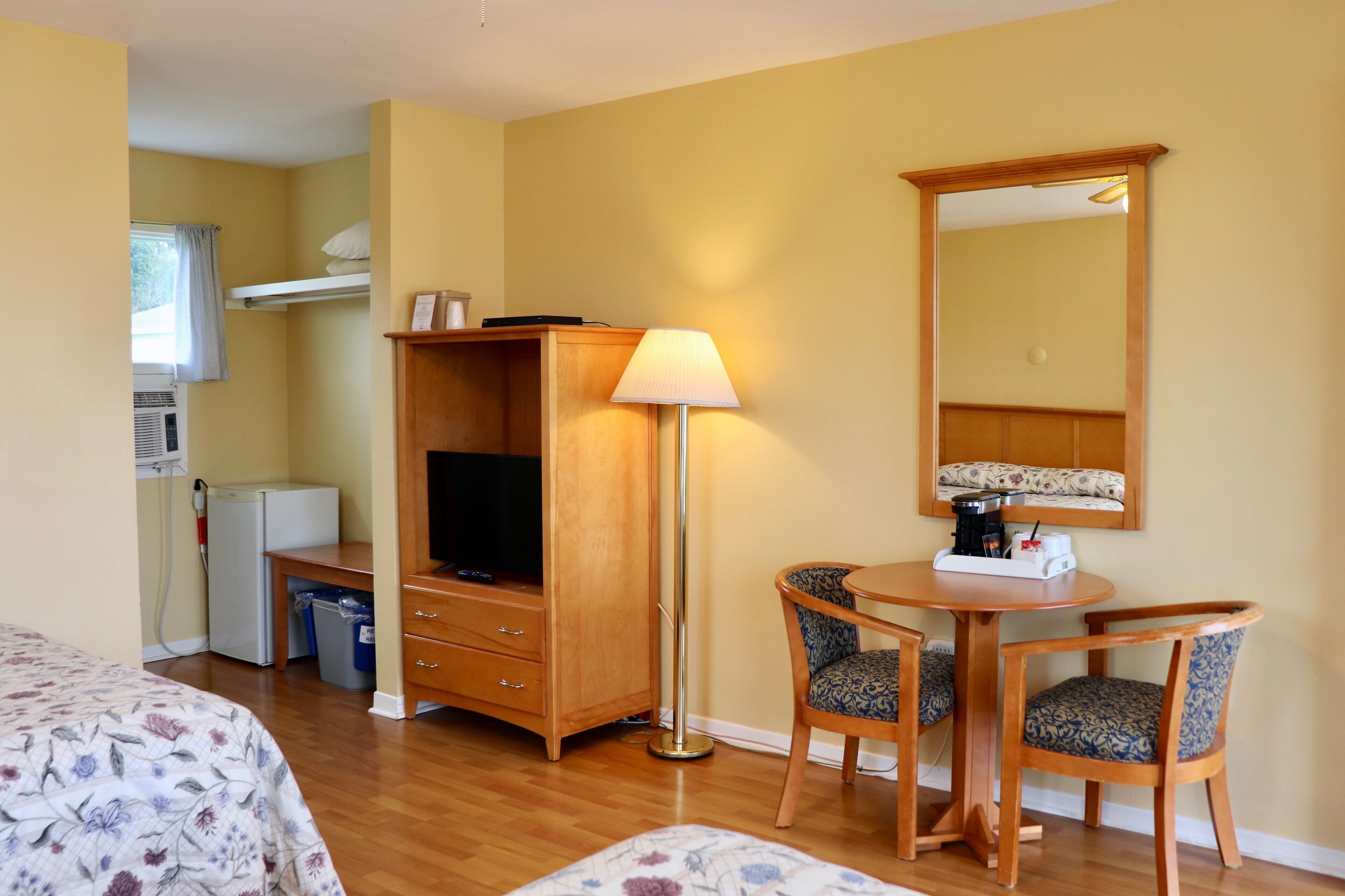 A hotel room with a bed, a desk with a TV, a lamp, a mirror, and a small kitchenette.