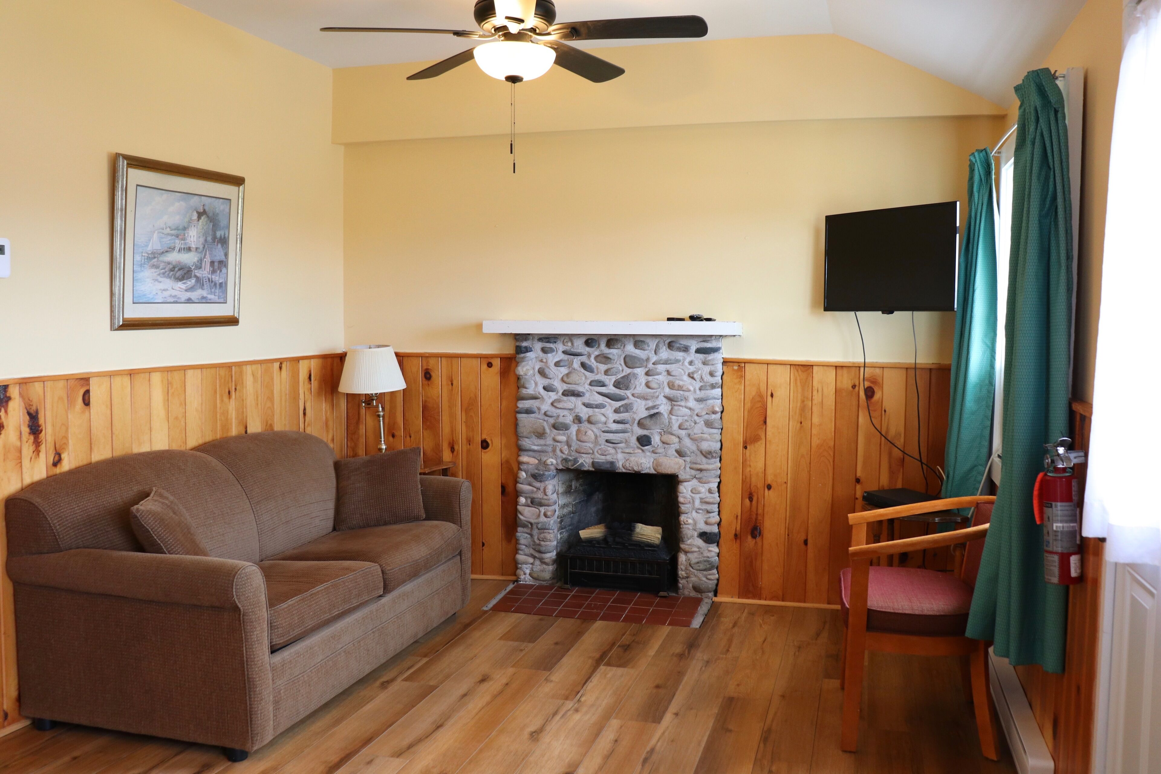 A cozy living room with a stone fireplace, wooden walls, a brown sofa, a television, and a ceiling fan.
