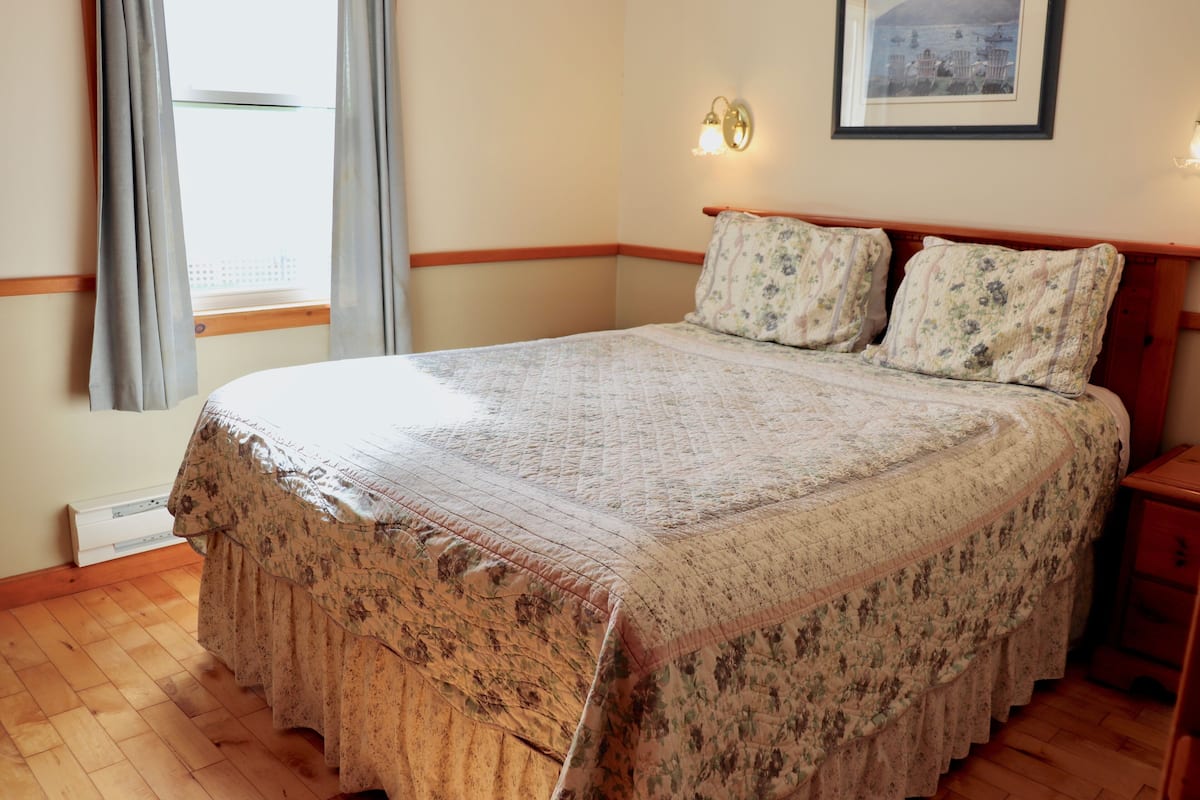 Una cama bien hecha con ropa de cama floral, una mesita de noche, una lámpara de pared y una ventana con cortinas.