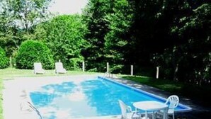 Una piscina rectangular rodeada por una cerca blanca, una pequeña mesa blanca con dos sillas, y algunas sillas de descanso al fondo.