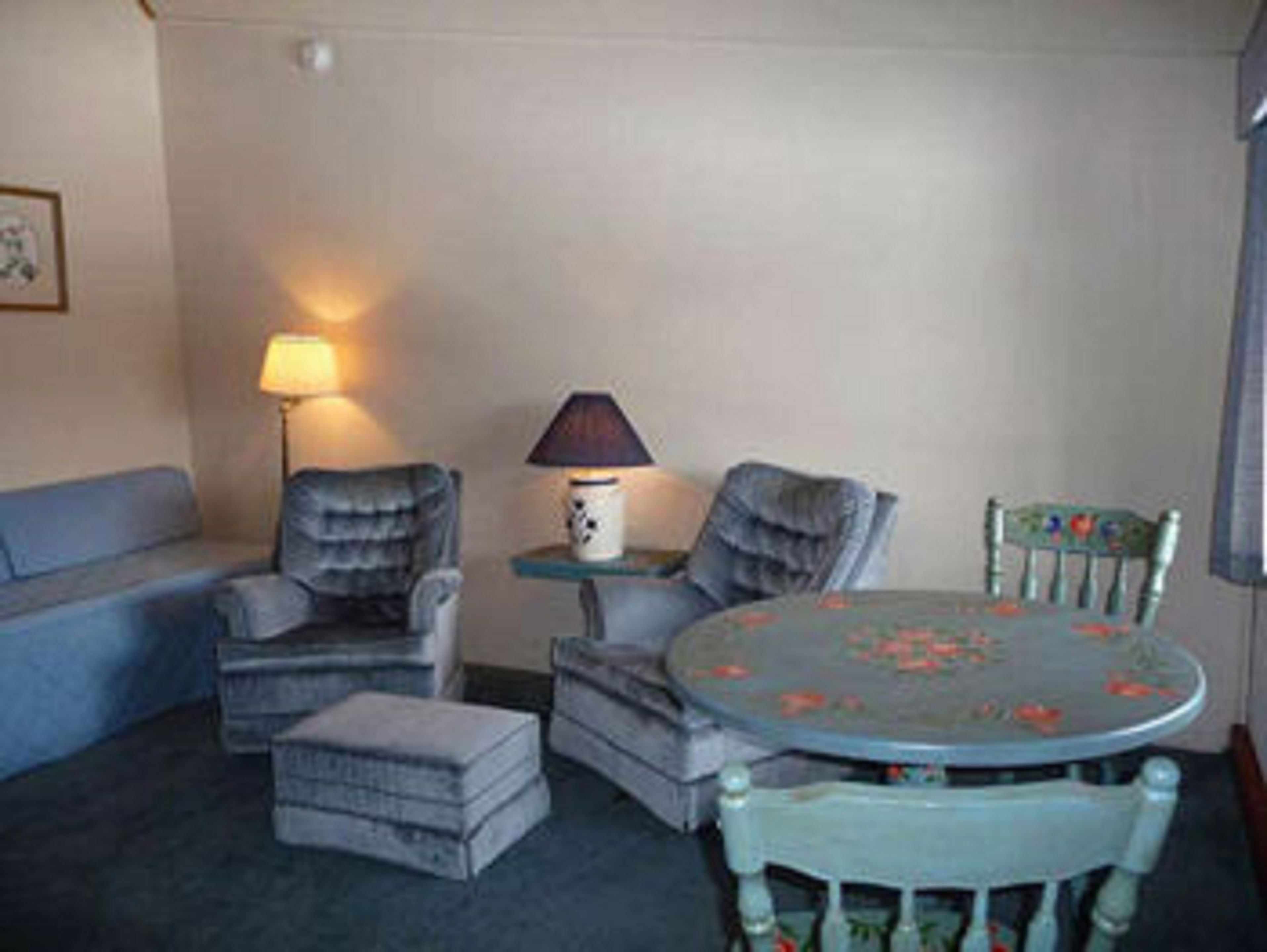 Ein gemütliches Zimmer mit einem blauen Sofa, zwei Sesseln, einem kleinen Tisch mit Lampe und einem runden Tisch mit Blumenmuster-Tischdecke.