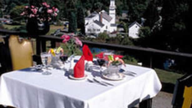 Mesa de comedor al aire libre con mantel blanco, un jarrón con flores y una iglesia y colinas al fondo.