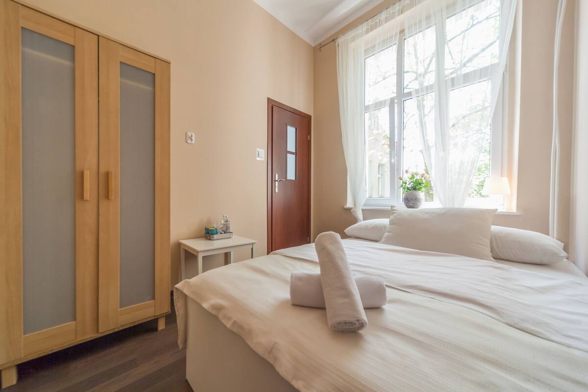 A bedroom with a bed, a wardrobe, a small table, and a window with curtains.