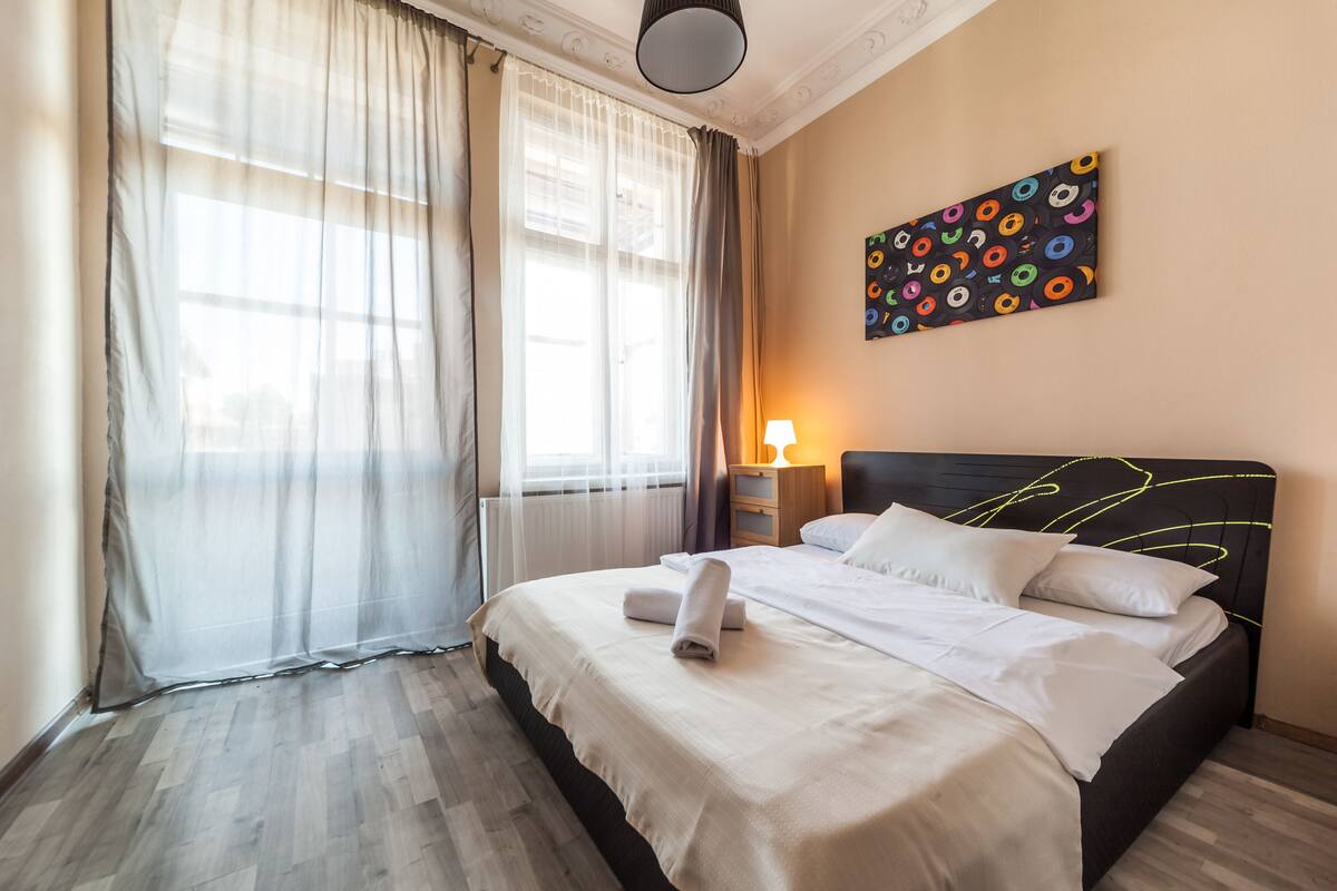 A hotel room with a bed, a window with sheer curtains, a wall art piece, and a bedside lamp.