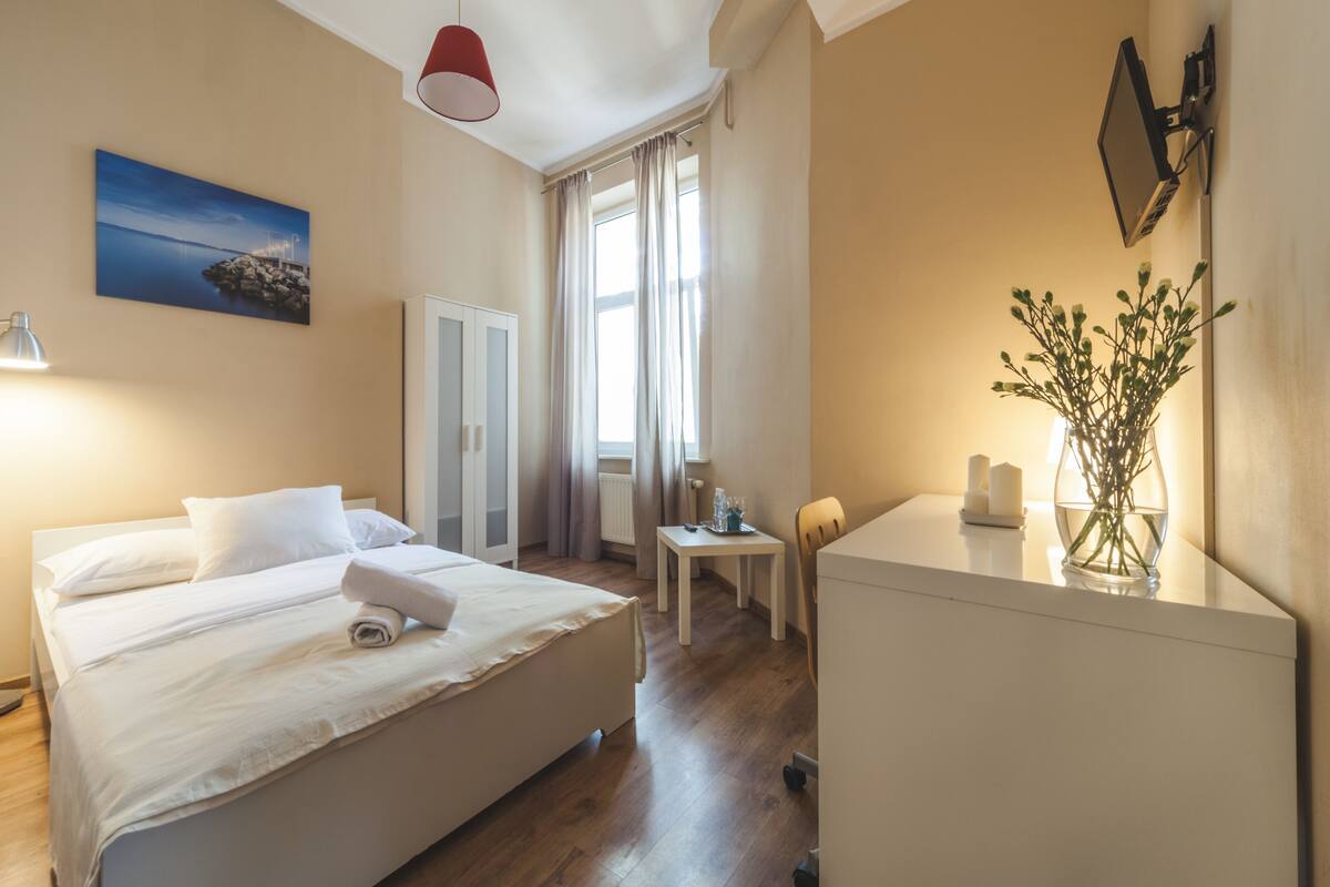 A hotel room with a bed, bedside table, desk, chair, lamp, mirror, and a painting on the wall.
