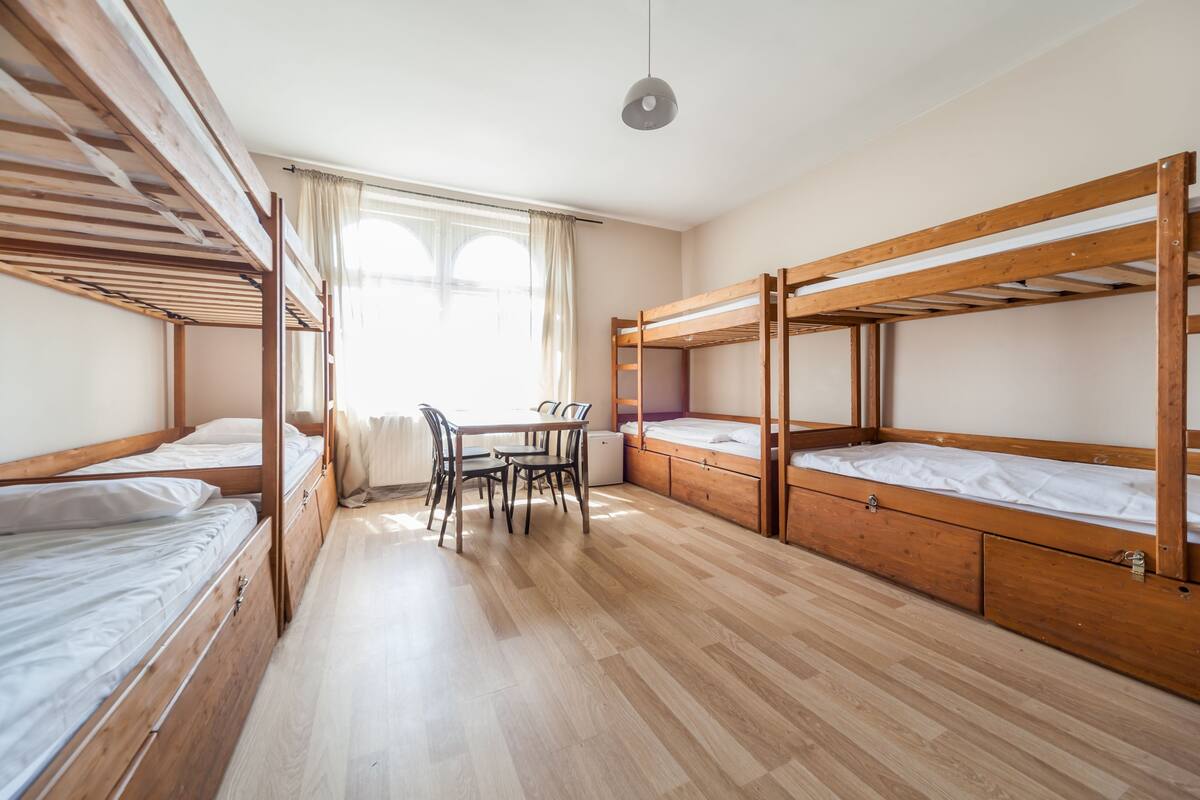 A dormitory room with bunk beds, a small table with chairs, and a window with curtains.
