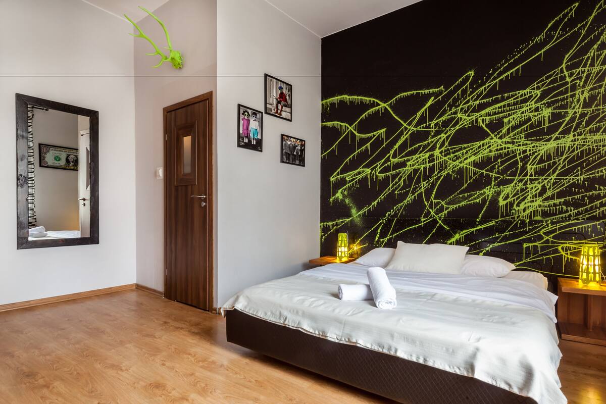 A bedroom with a bed, a wooden door, a mirror, and a wall with a green abstract design.