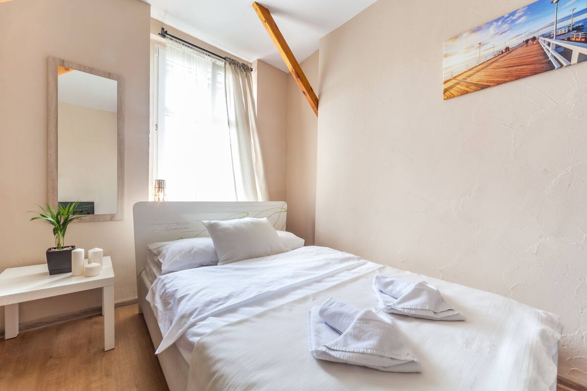 A bedroom with a bed, a bedside table with a plant, a mirror, and a painting on the wall.