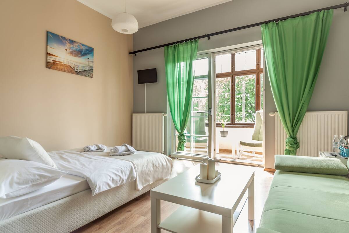 A room with a bed, a green sofa, a table, a painting, and a view of the outdoors.
