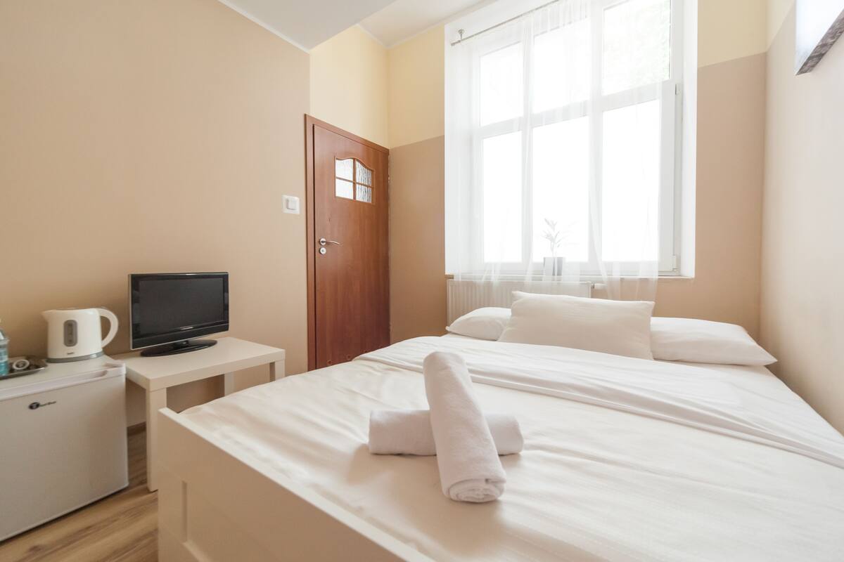 A hotel room with a bed, a TV, a small table with a kettle, and a wardrobe.