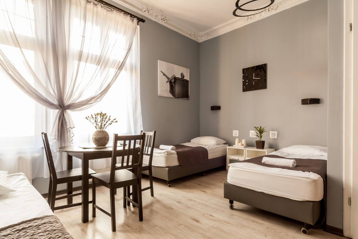 A modern bedroom with a wooden dining table, two single beds, a small bedside table with a plant, and a wall clock.