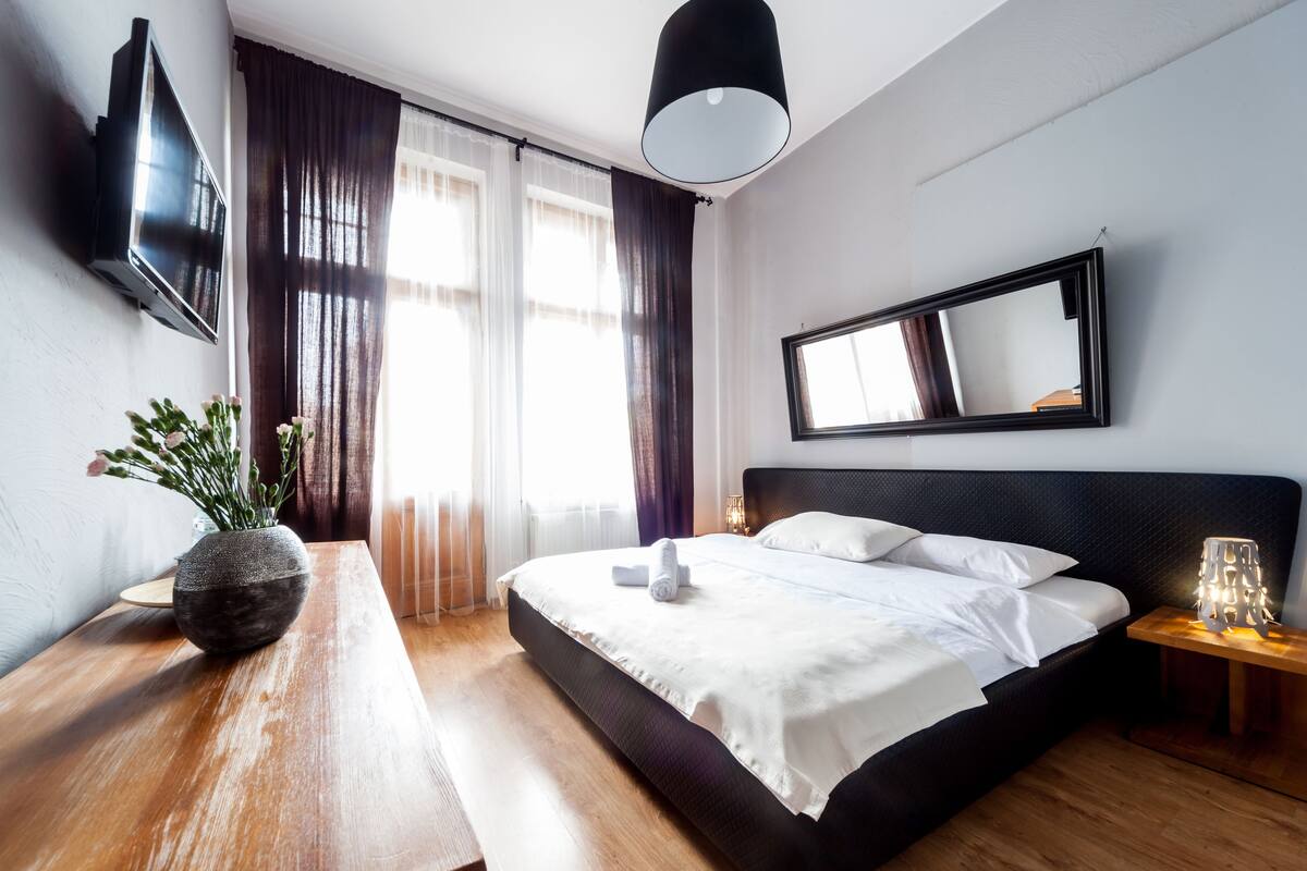 A modern hotel room with a large bed, a flat-screen TV mounted on the wall, a bedside table with a lamp, and a window with curtains.