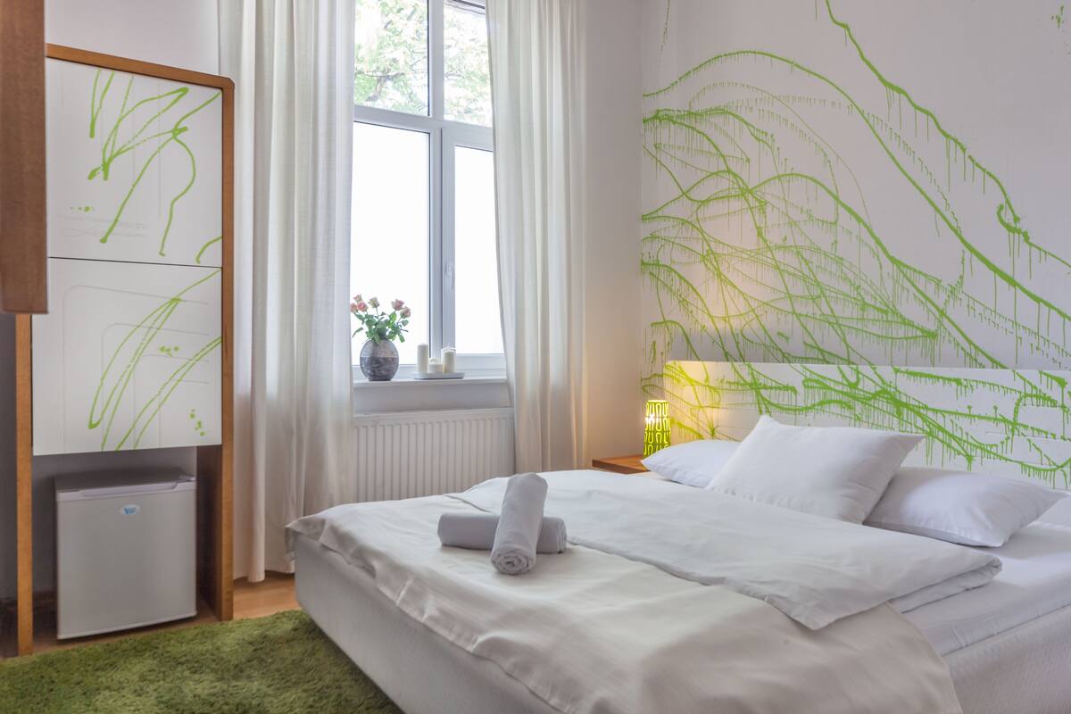 A bedroom with a bed, a nightstand, a lamp, a window with curtains, and a wall with green abstract art.