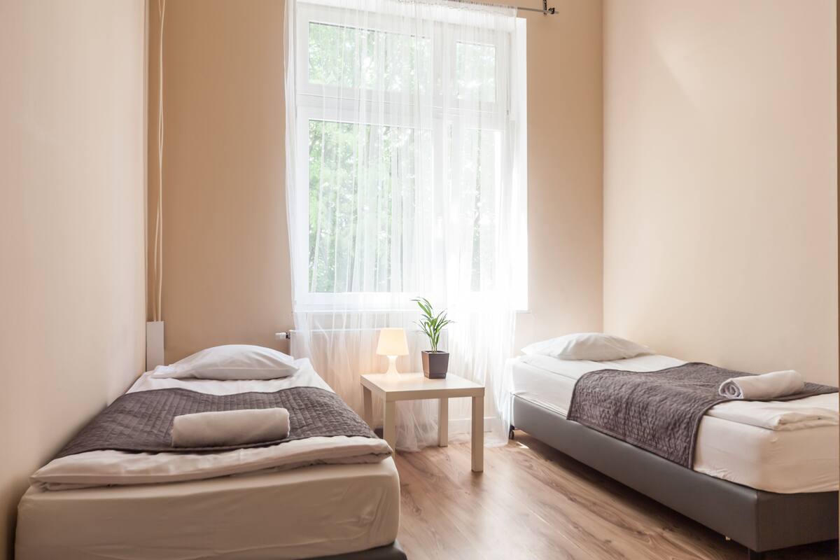 A small, well-lit bedroom with two beds, a bedside table with a lamp, and a window with sheer curtains.