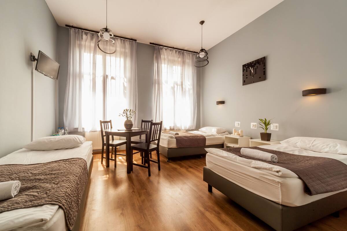 A modern hotel room with two beds, a dining table, and a small kitchen area.