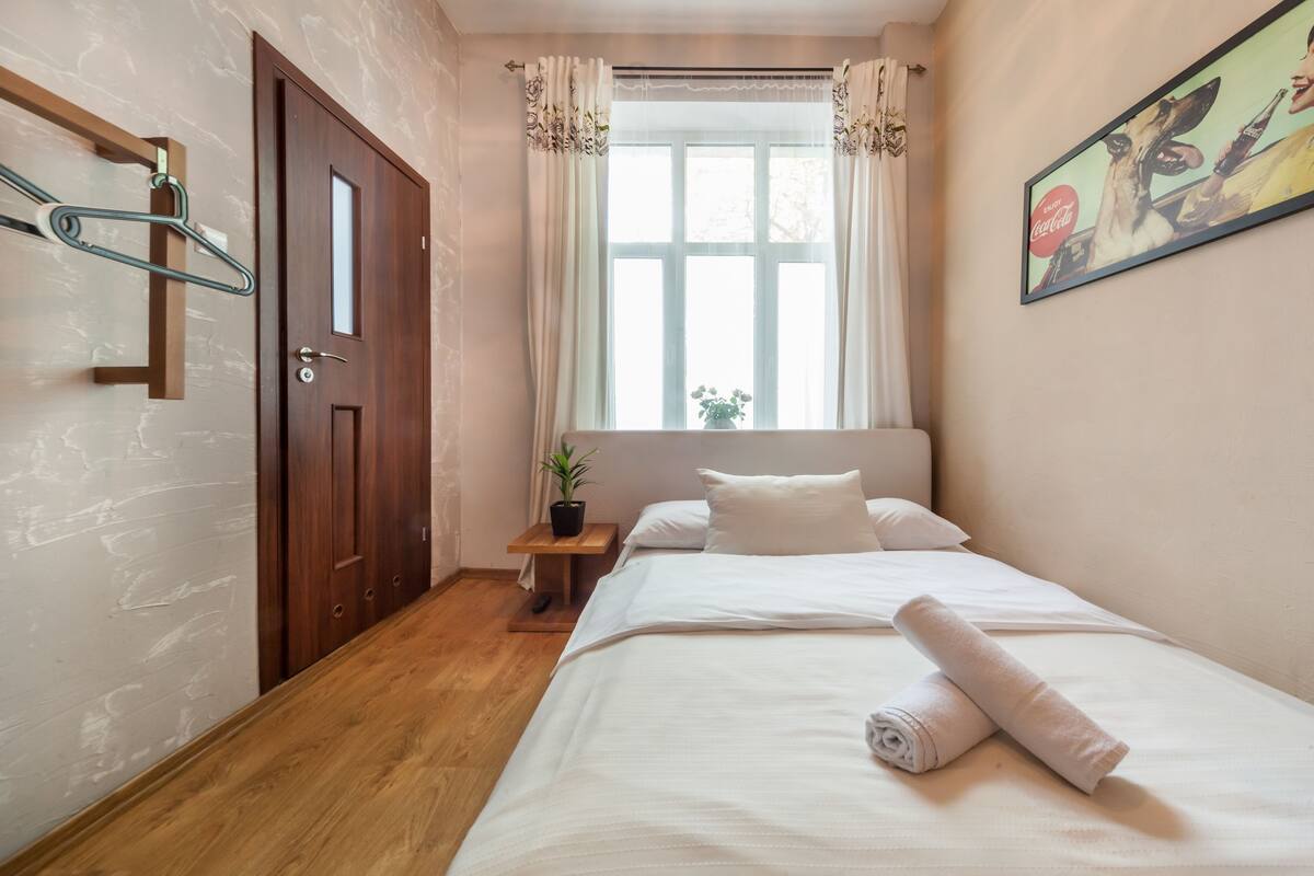 A bedroom with a wooden door, a window with curtains, a bed with white bedding, and a small table with a plant.