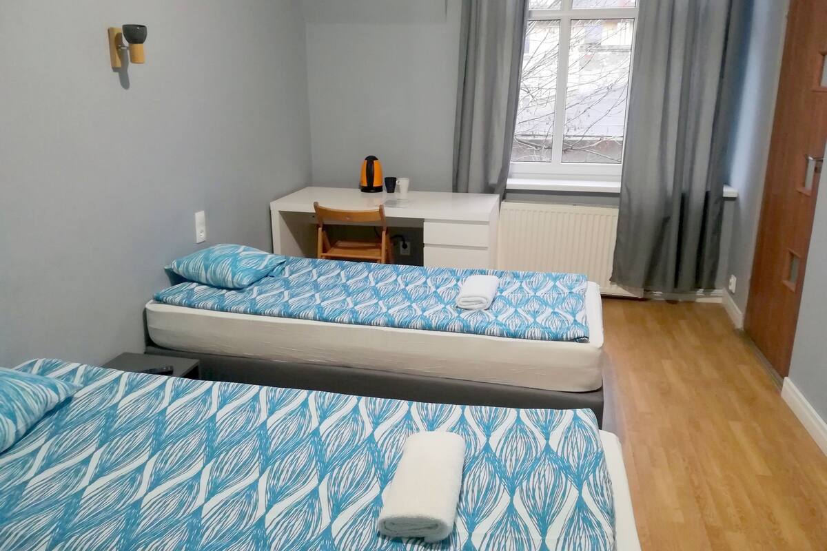 A small, tidy room with two beds, a desk, and a window with curtains.