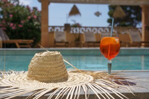 A straw hat and a glass of drink on a table by a pool with lounge chairs and a thatched umbrella in the background. - Apartamentos Andreas (Ses Salines)