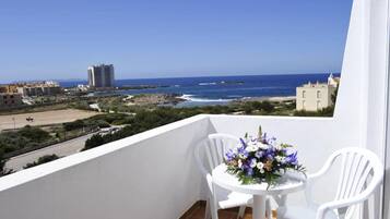 A balcony with two white chairs and a table, overlooking a beach and ocean with buildings in the background.