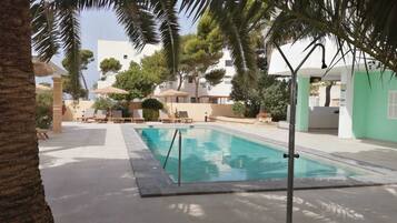 A swimming pool with lounge chairs and umbrellas, surrounded by palm trees and buildings.
