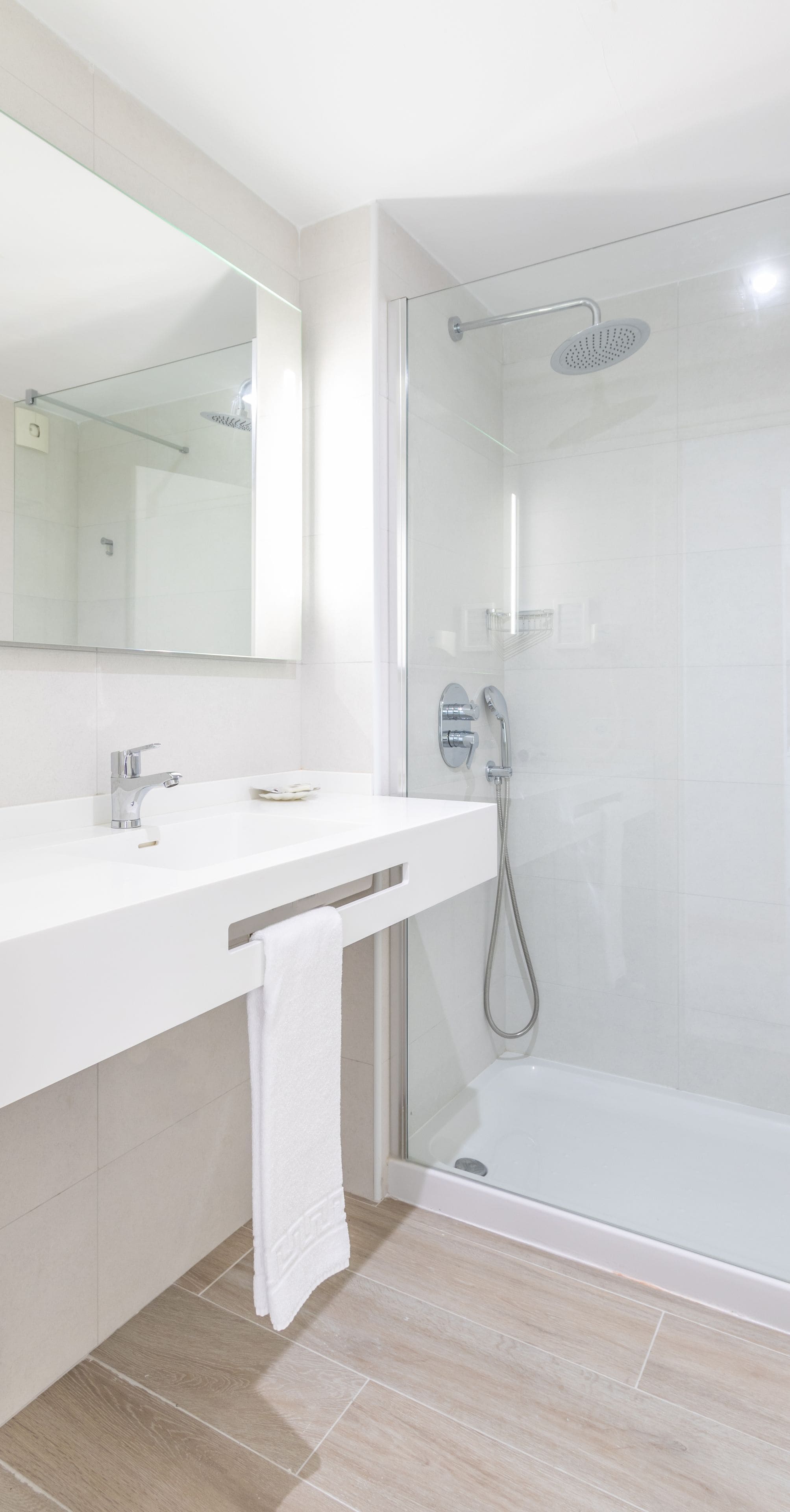 Un baño moderno con ducha envuelta en vidrio, un tocador blanco con espejo y una toalla colgada en un perchero.