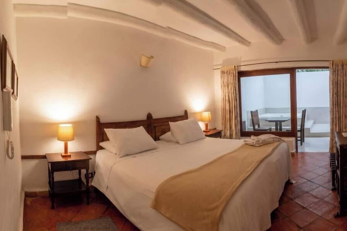 Een hotelkamer met een bed, twee bedlampen, een houten hoofdbord, een tafeltje en uitzicht naar buiten door een glazen deur.