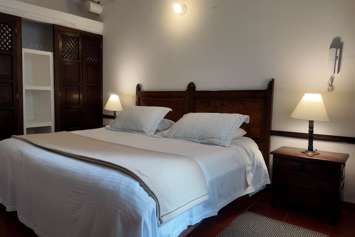 Een keurig opgemaakt bed met witte lakens, een houten hoofdbord, een bedlampje en een houten garderobe met verstelbare deuren.