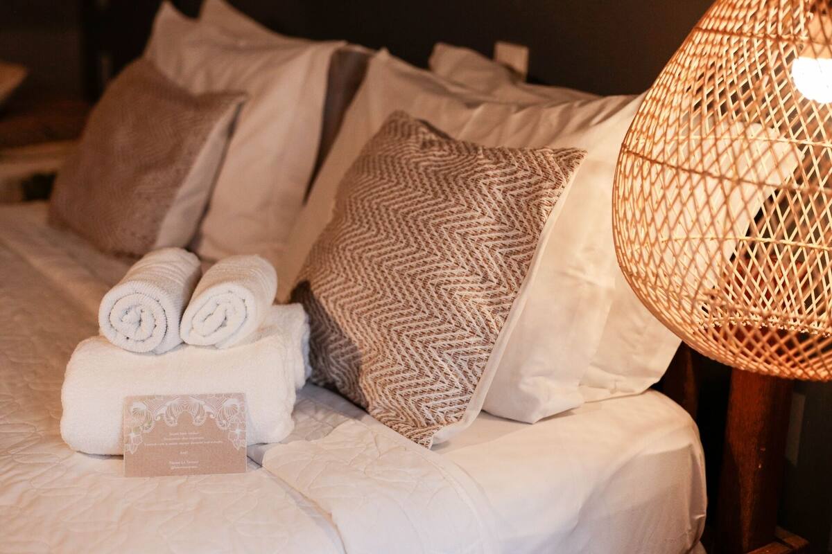 Un lit bien fait, avec des serviettes pliées, un coussin décoratif, une petite carte et une lampe en osier.