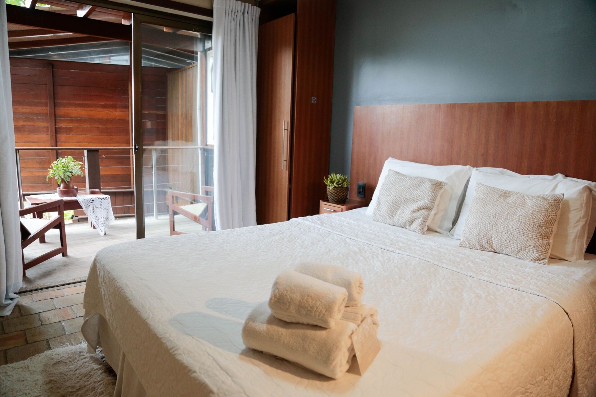 A bedroom with a large bed, a wooden headboard, a balcony with a chair, and a potted plant.