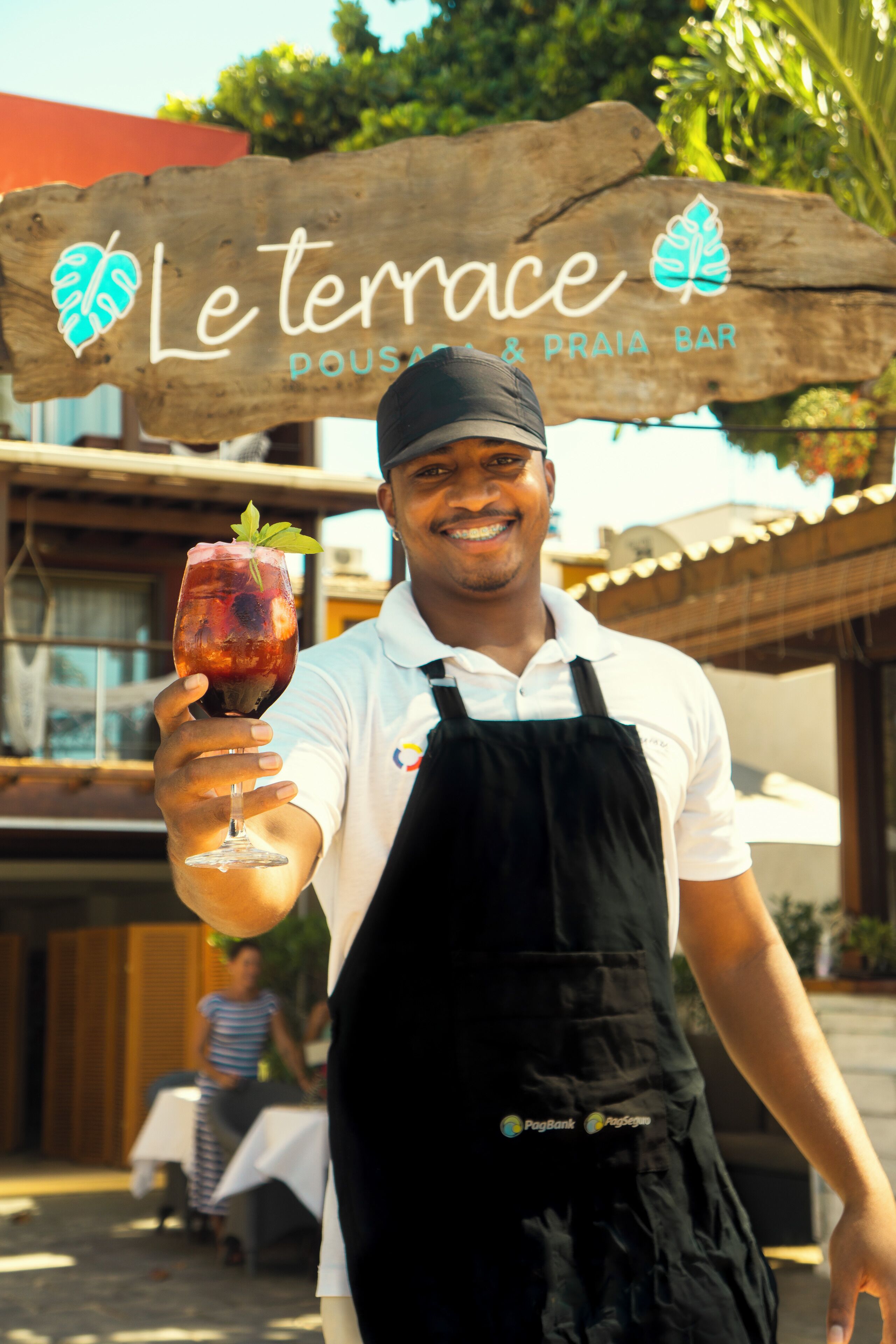 A person holding a glass of drink in front of a wooden sign that reads "Le terrace DOUAS & PRAIA BAR".