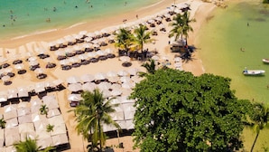 Vue aérienne d’un complexe hôtelier en bord de mer, avec des rangées de villas situées au bord de la plage, un espace piscine et des eaux turquoise cristallines.