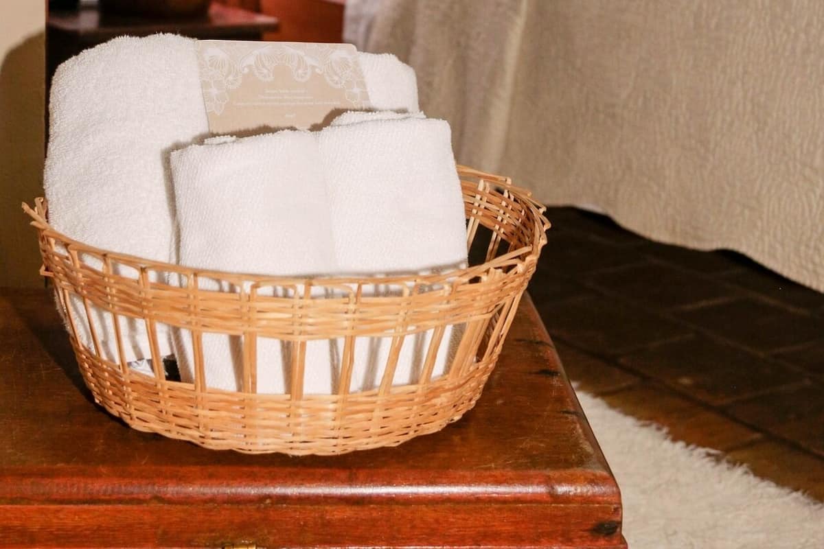 Un panier en osier contenant des serviettes pliées, posé sur une table en bois dans une chambre à coucher équipée d’un lit, d’oreillers et d’une table de chevet avec une lampe.