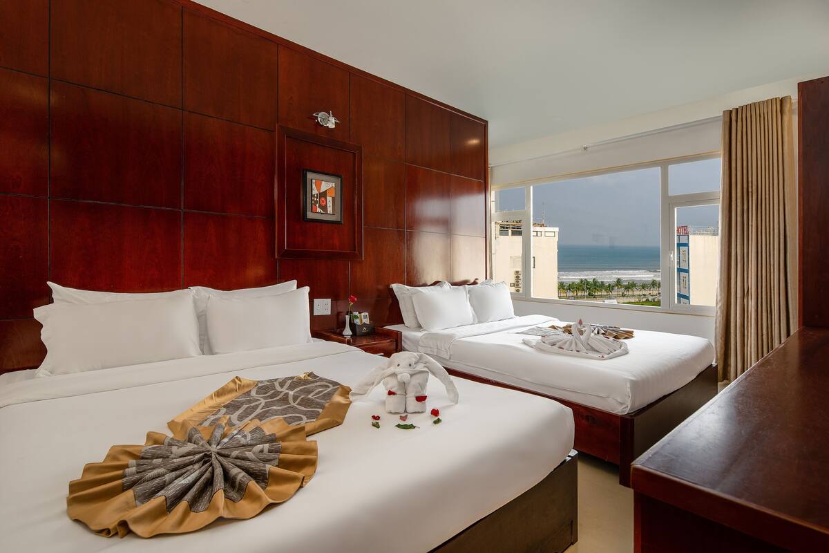 A hotel room with two beds, a large window with a view of the sea, and a wooden headboard.