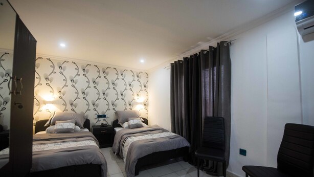 A bedroom with two beds, a nightstand, a chair, and a patterned wallpaper. - Niagara Inn (Accra)