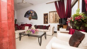 A living room with a ceiling fan, a sofa, a coffee table with flowers, a TV, and a window with curtains. - Niagara Inn (Accra)
