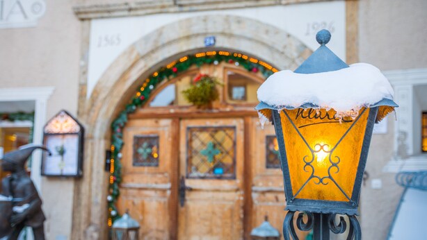 A street lamp with snow on top, a statue, and a building with an archway and decorations. - Hotel Klarer (Zuoz)