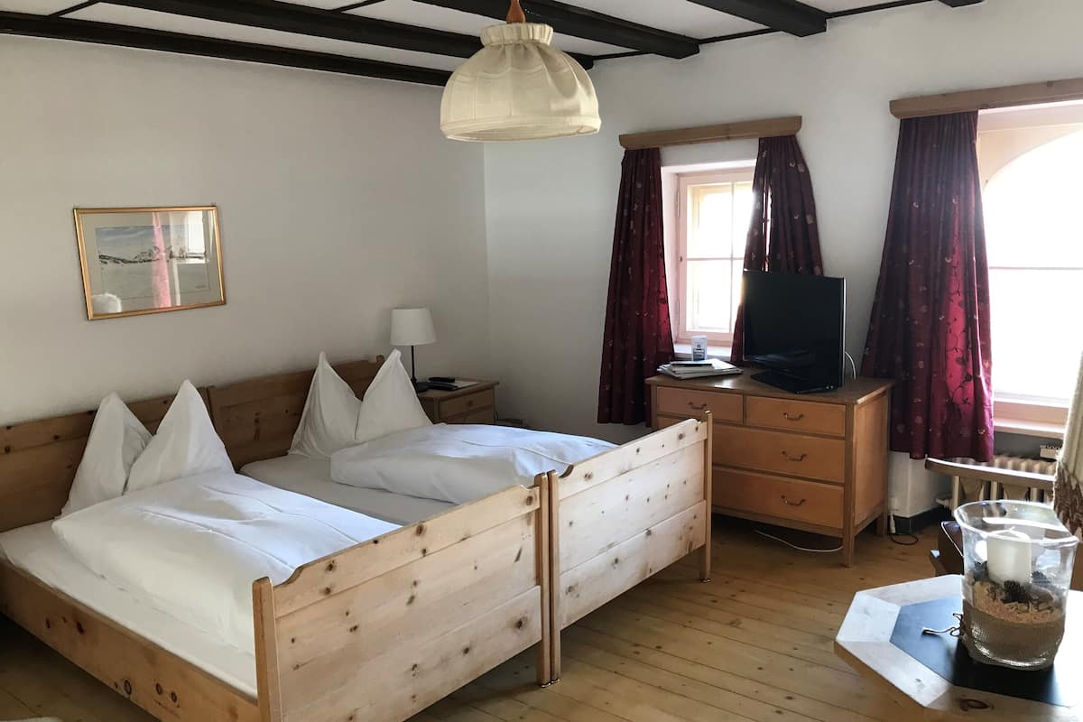 Una cama de madera con ropa blanca de cama, una mesita de noche con una lámpara, un televisor en un aparador y una ventana con cortinas.