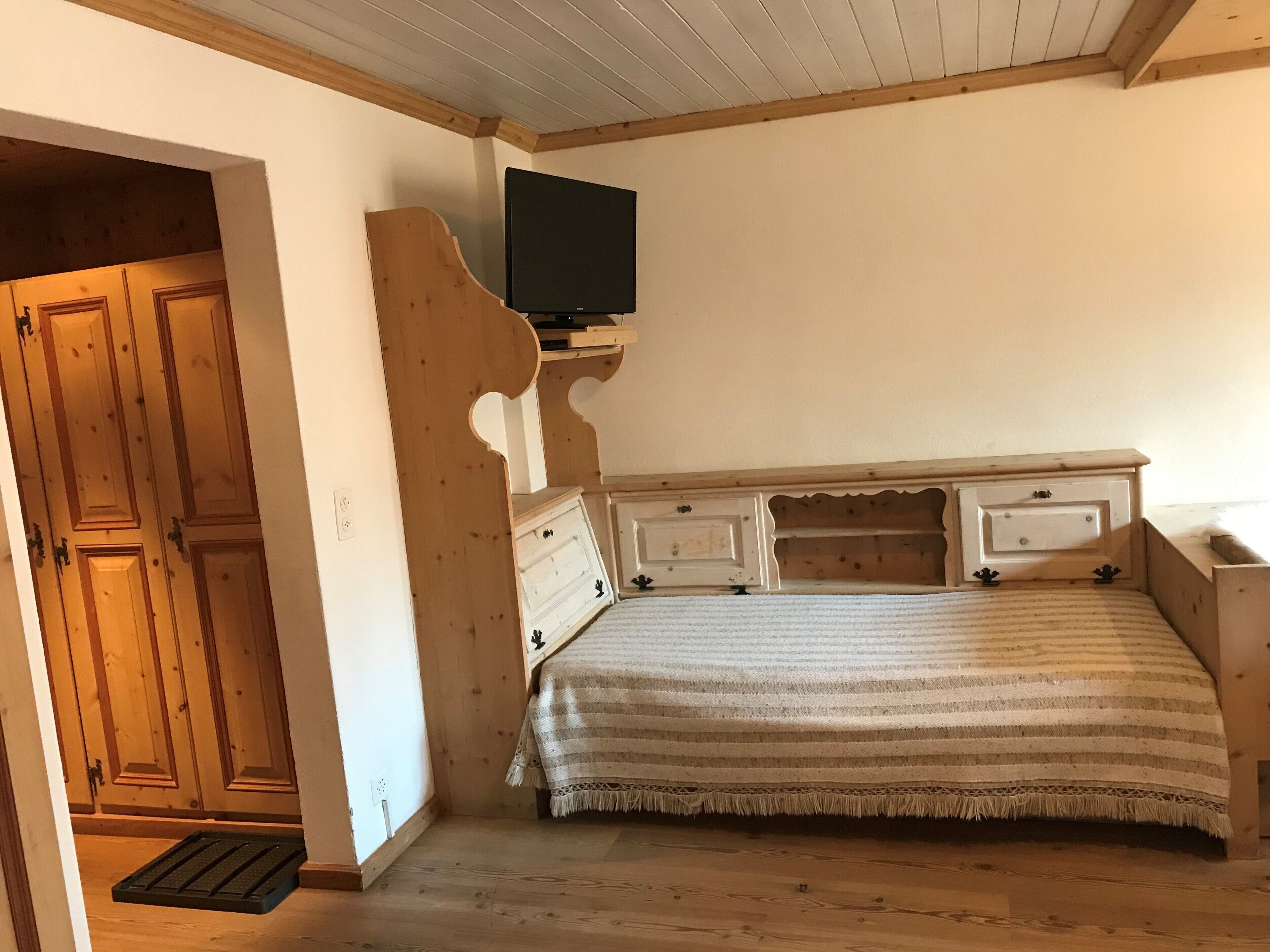 A bedroom with a wooden bed, a television mounted on the wall, and a wooden wardrobe.