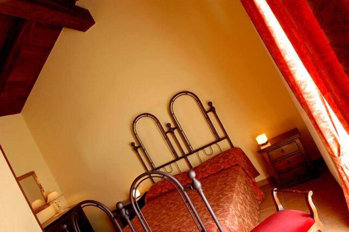 Een slaapkamer met een metalen bed, een houten stoel, een nachtkastje met een lamp en een raam met gordijnen.