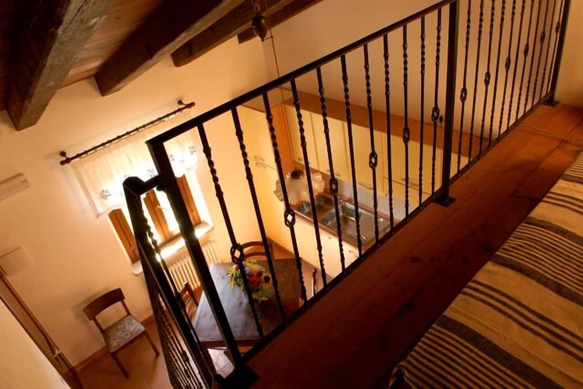 Een trap met een houten leuning en zwarte spijbels, die leidt naar een kamer met een bed, een stoel en een tafel met een bloevas.