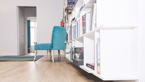 Una habitación con piso de madera, una silla color turquesa y una estantería blanca llena de libros.