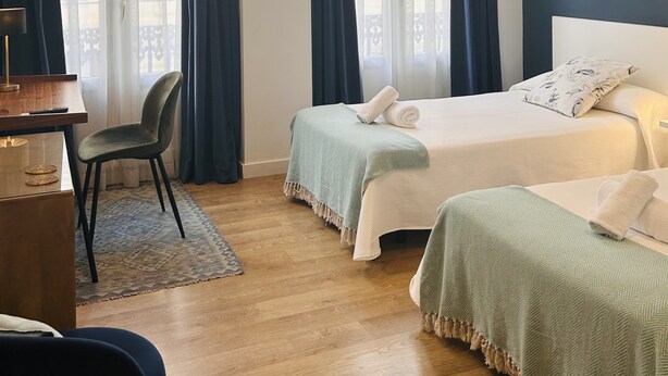 Habitación de hotel con dos camas, un escritorio, una silla y ventanas amplias con cortinas.