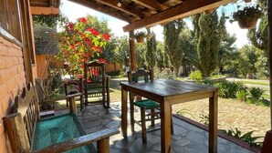 Un patio al aire libre con una mesa y bancos de madera, rodeado de follaje y una pared de ladrillo.