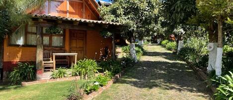 A wooden cabin with a porch, surrounded by a garden with various plants and a cobblestone path leading to the entrance.