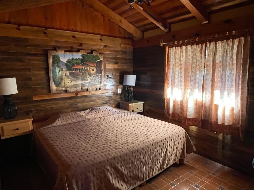 Een slaapkamer van een houten hutje, met een bed, twee nachtkastjes, een lamp, een schilderij aan de muur en een raam met gordijnen.