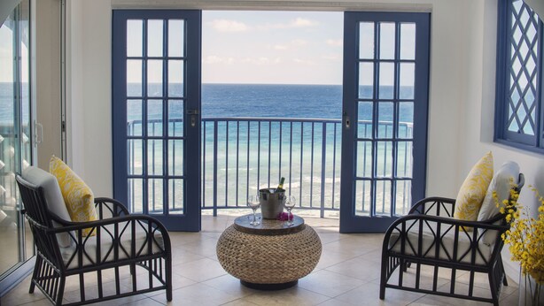 Un balcon avec deux fauteuils en osier, une table ronde avec une bouteille et des verres, et une vue sur l’océan.